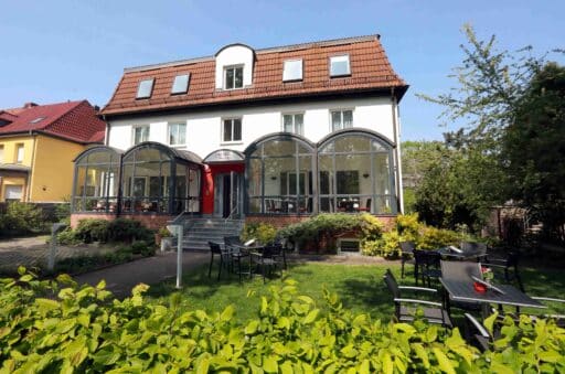 Blick auf das Hotel mit Grünfläche davor. Weißes Gebäude mit rotem Dach, davor ein Wintergarten in dem Tische und Stühle zu sehen sind. Davor ist eine Wiese auf verteilt Tische und Stühle stehen.
