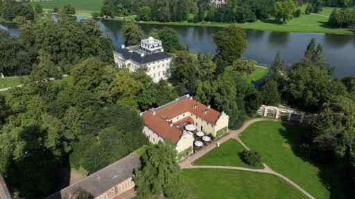 Luftaufnahme. Zwei Gebäude sind zu sehen. Vorne ein gelbes Haus und dahinter ein weißes Gebäude, Schloss Wörlitz. Im Hintergrund ist ein See.