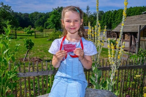 Ein blondes Mädchen mit blauem Kleid hält eine Gästecard der WelterbeRegion in die Kamera.