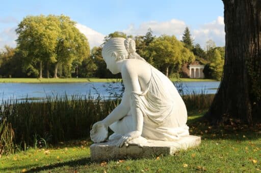 Eine Statur aus Stein sitzt auf dem Boden. Es ist eine Frau, eine Nymphe. In der einen Hand hält sie eine Muschel. Ihr Gesicht ist abgewendet. Im Hintergrund ist ein See zu erkennen.